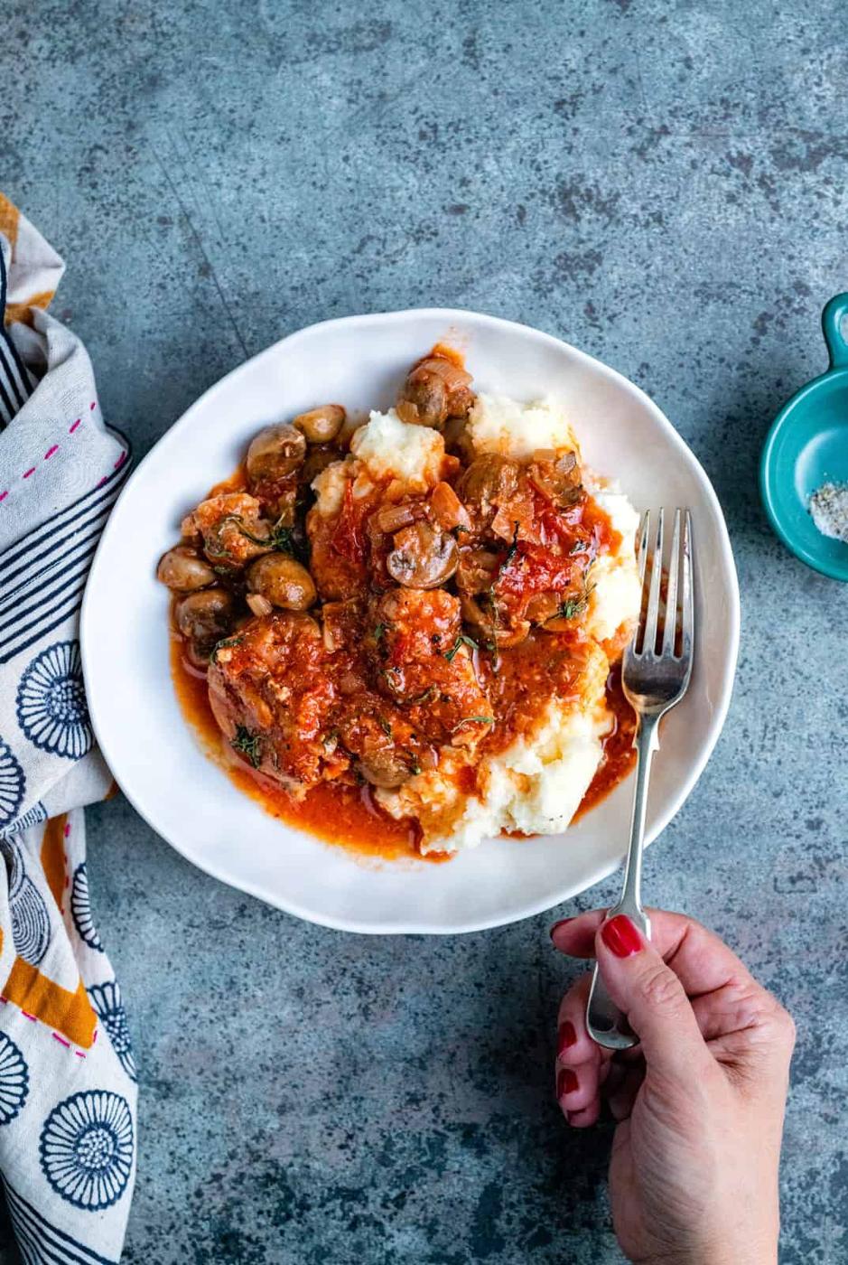A hand holding a fork is poised over a plate of mashed potatoes topped with Slow Cooker Chicken Chasseur—a tomato-based stew with mushrooms and herbs. A patterned cloth and a small bowl of salt sit nearby on a textured blue surface.