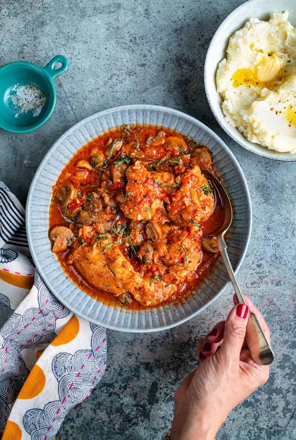 A hand holding a spoon over a bowl of Slow Cooker Chicken Chasseur with vegetables in a tomato-based sauce. Next to it is a bowl of mashed potatoes with butter, a small blue bowl with salt, and a colorful napkin.