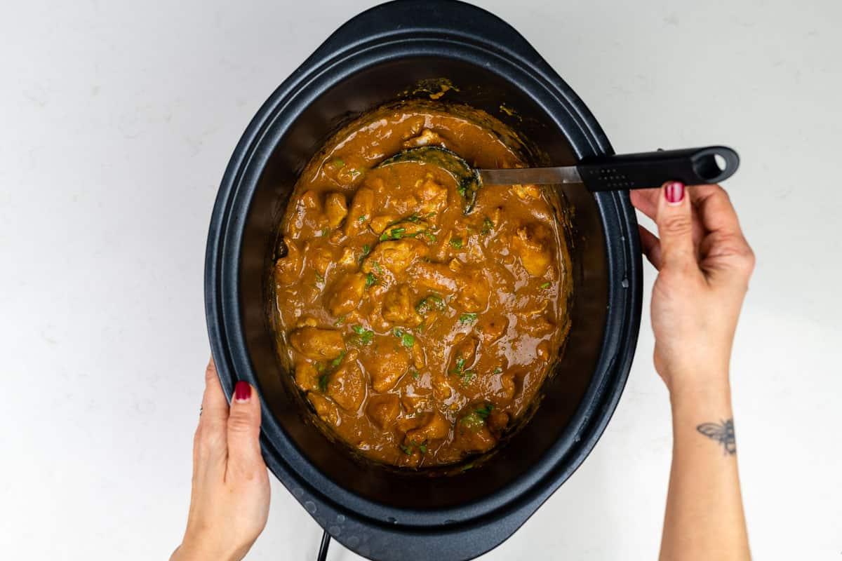 A person with red nail polish and a small wrist tattoo stirs a thick, brown Slow Cooker Chicken Curry with chunks of meat and herbs in a black slow cooker against a white background, showcasing an easy curry recipe.