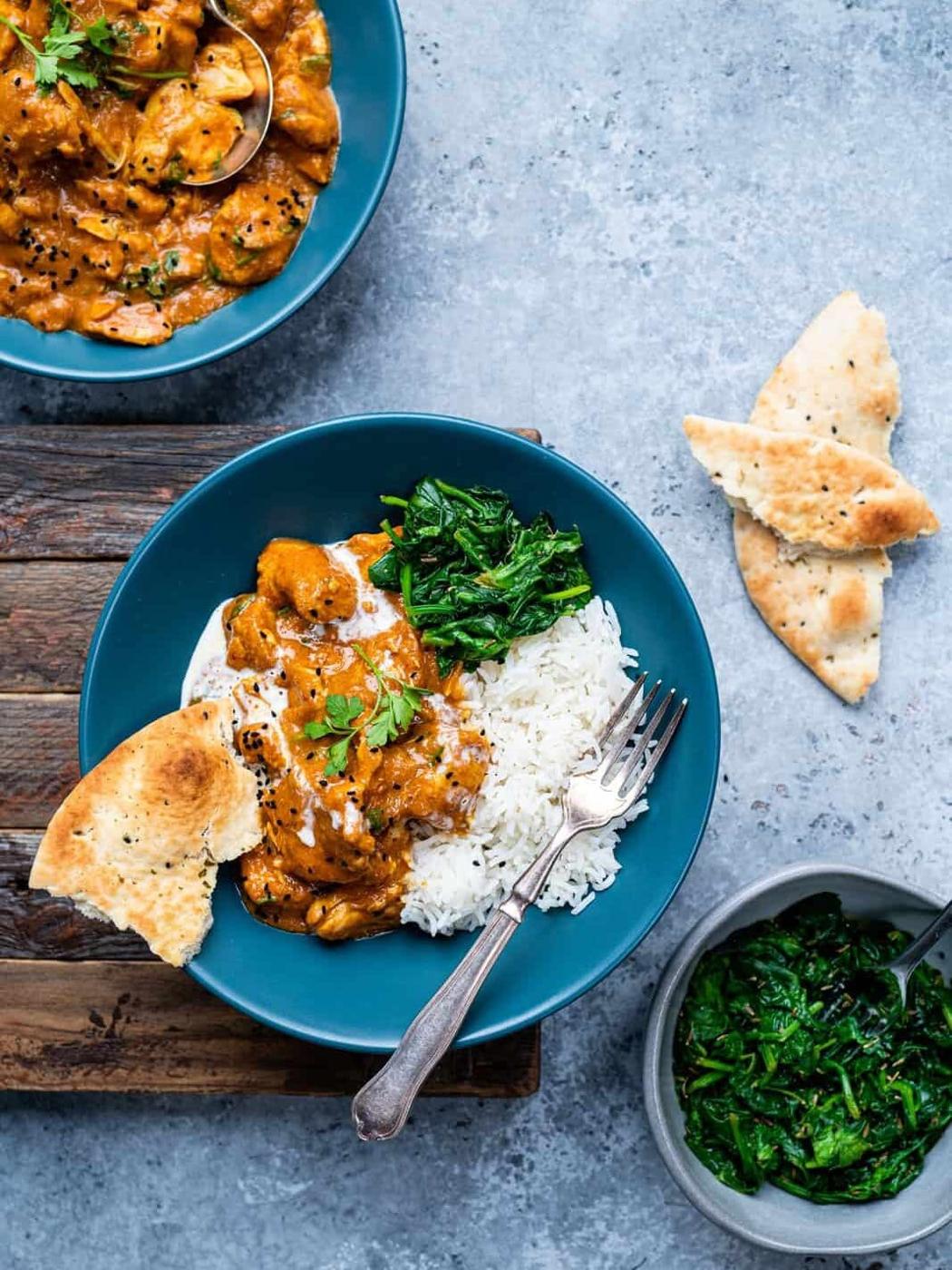 A blue bowl filled with rice, creamy easy curry recipe, sautéed spinach, and naan bread, with a fork on the side. Another bowl, naan pieces, and a small bowl of spinach are nearby on a gray background.
