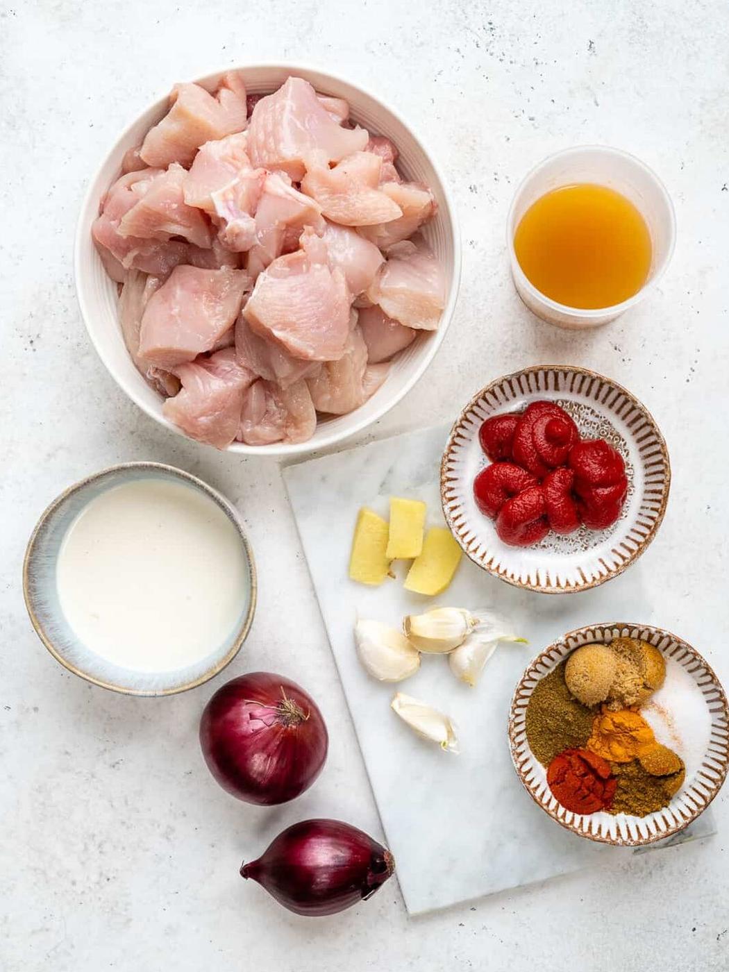 Top-down view of Slow Cooker Chicken Curry ingredients: raw diced chicken, broth, cream, spices, tomato paste, ginger, garlic cloves, and whole red onions—everything you need for an easy curry recipe all laid out on a white surface.