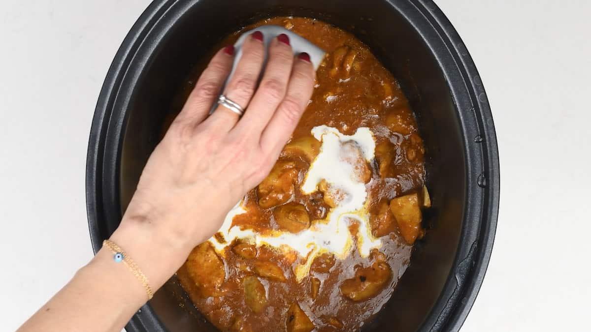 A hand with red nail polish pours cream into a slow cooker filled with a rich brown curry containing potatoes and mushrooms—an easy curry recipe perfect for making Slow Cooker Chicken Curry at home.