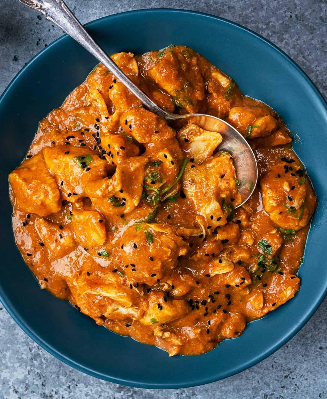 A blue bowl filled with creamy orange Slow Cooker Chicken Curry, garnished with black sesame seeds and herbs, with a silver spoon resting inside—a perfect easy curry recipe for any occasion.