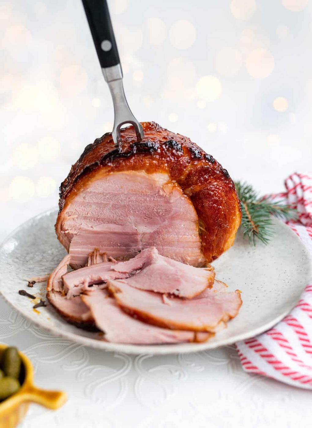 A slow cooker gammon glazed ham with several slices cut and fanned out on a white plate, garnished with a small pine sprig. A carving fork is inserted into the top of the ham, and a red and white striped towel sits in the background.