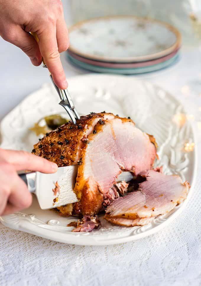 A person slices a glazed, cooked slow cooker gammon on a white platter with a carving fork and knife. Slices of ham are fanned out on the plate, and stacked plates are visible in the background.