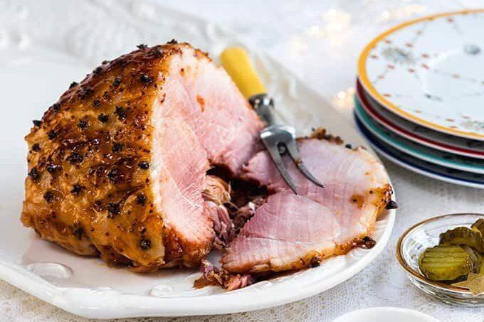 A glazed ham studded with cloves is partially sliced on a white platter, with a carving fork resting beside it. Stacked plates and a small dish of pickles are in the background.