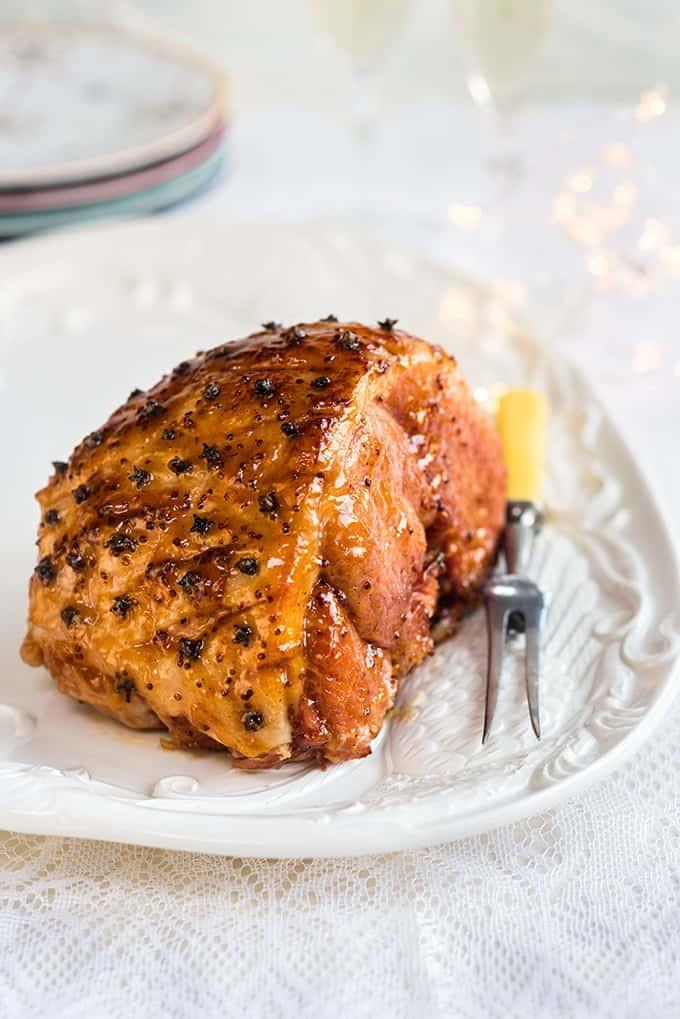A glazed, baked slow cooker gammon studded with cloves sits on a white platter with a carving fork, ready to be sliced. The background features stacked plates and soft, festive lighting.