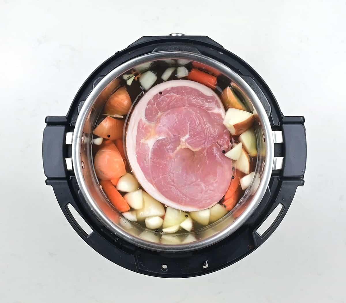 An overhead view of an Instant Pot filled with a large cut of raw ham, chopped onions, carrots, and seasonings submerged in liquid – perfect for making slow cooker gammon, ready to be cooked.