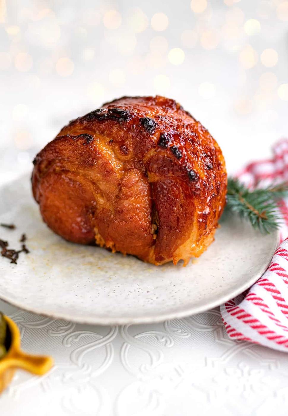 A glazed, roasted slow cooker gammon sits on a white plate, garnished with a small pine branch. The background is softly blurred with warm lights, and a red-and-white striped cloth is nearby.
