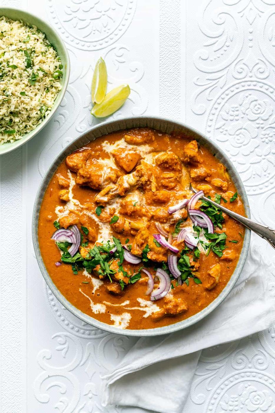 A bowl of creamy orange-colored Slow Cooker butter chicken, garnished with sliced red onions, fresh herbs, and a swirl of cream, sits on a white patterned surface next to lemon wedges and a bowl of rice with chopped herbs.