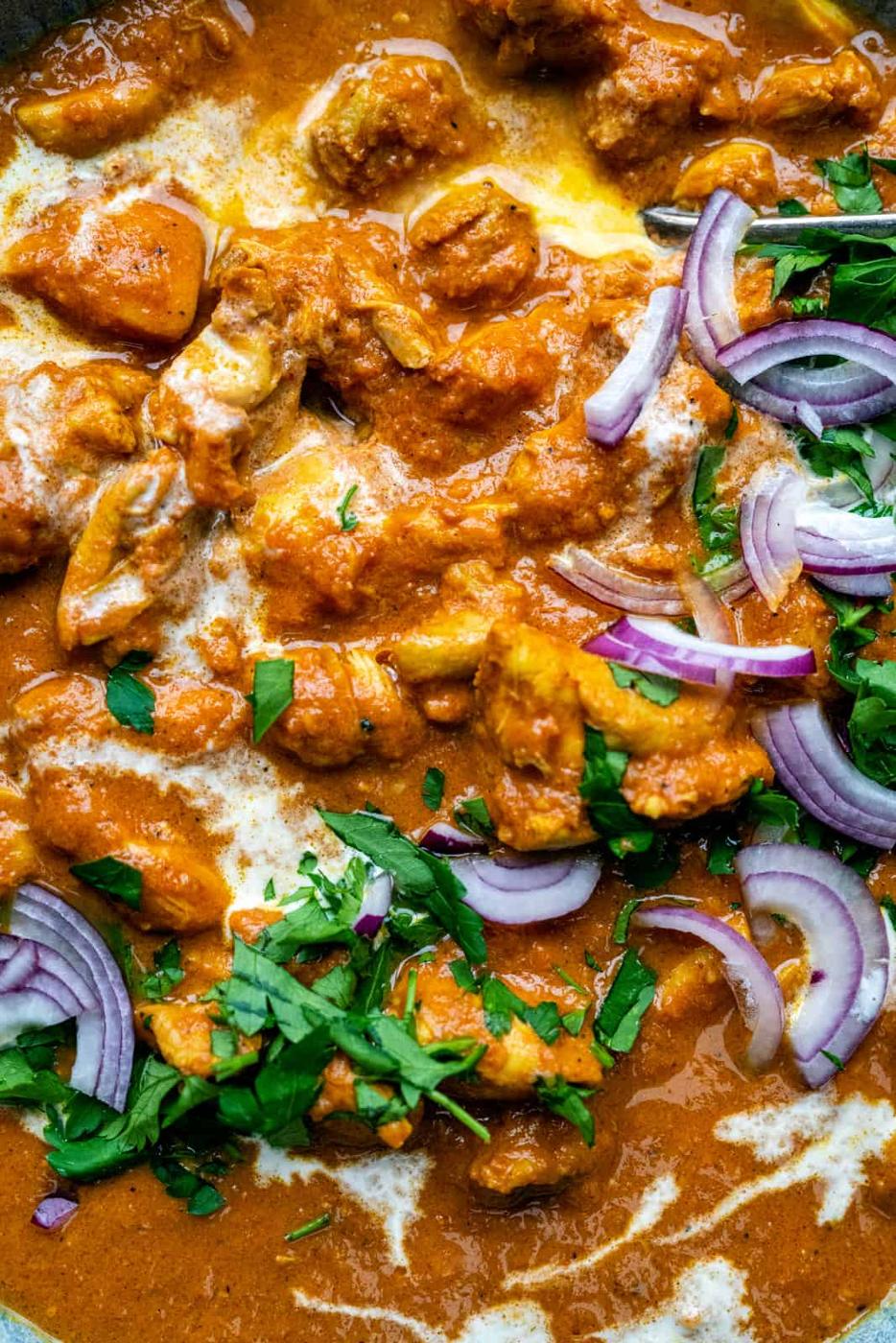 Close-up of creamy orange Butter Chicken with tender chunks of chicken, topped with fresh cilantro, slices of red onion, and a swirl of cream.