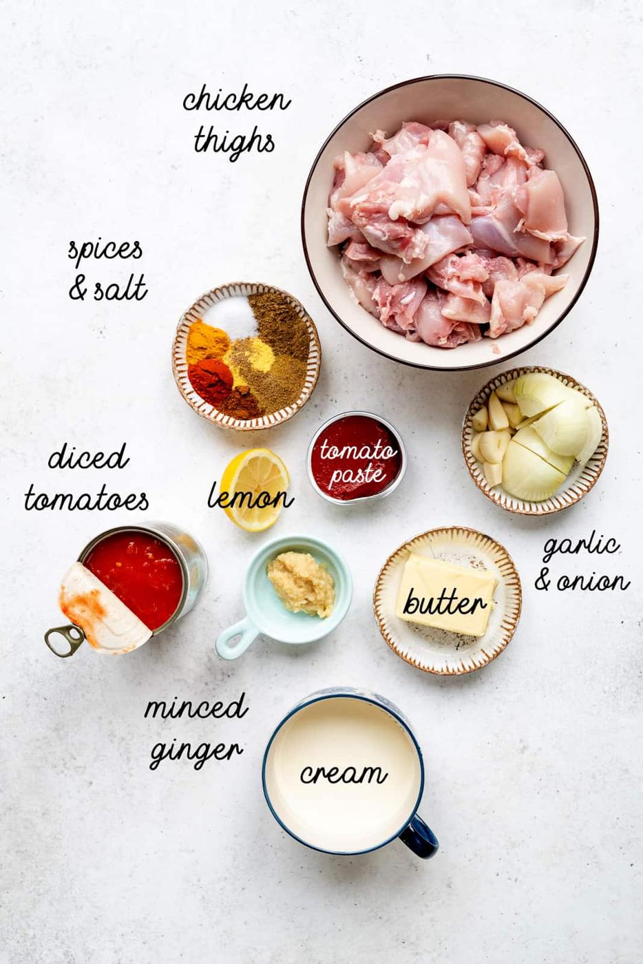 A flat lay of ingredients in bowls and cups, labeled: chicken thighs, spices & salt, diced tomatoes, lemon, tomato paste, butter, garlic & onion, minced ginger, and cream—perfect for making Slow Cooker Butter Chicken—on a white background.