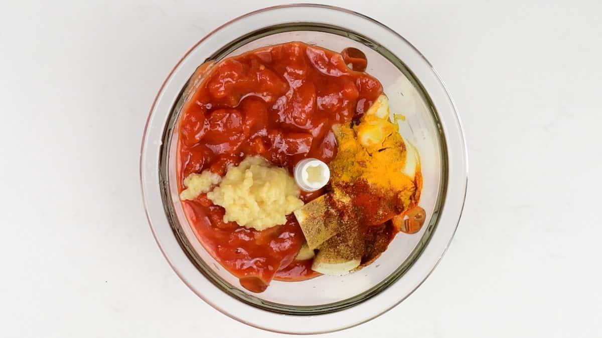 A glass bowl containing chopped tomatoes, minced garlic, sliced onions, turmeric, and chili powder—essential ingredients for a classic Butter Chicken—viewed from above on a white background.