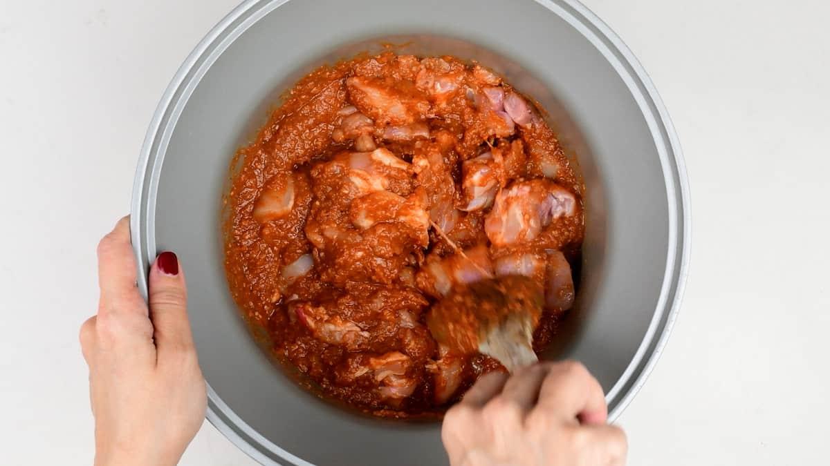A person stirs raw chicken pieces coated in a red marinade, preparing them for Butter Chicken, inside a grey mixing bowl using a spoon. The bowl sits on a white surface.