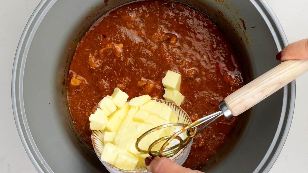 A person adds cubed butter from a small glass bowl into a pot of rich, red-brown Slow Cooker butter chicken sauce with chunks of meat.