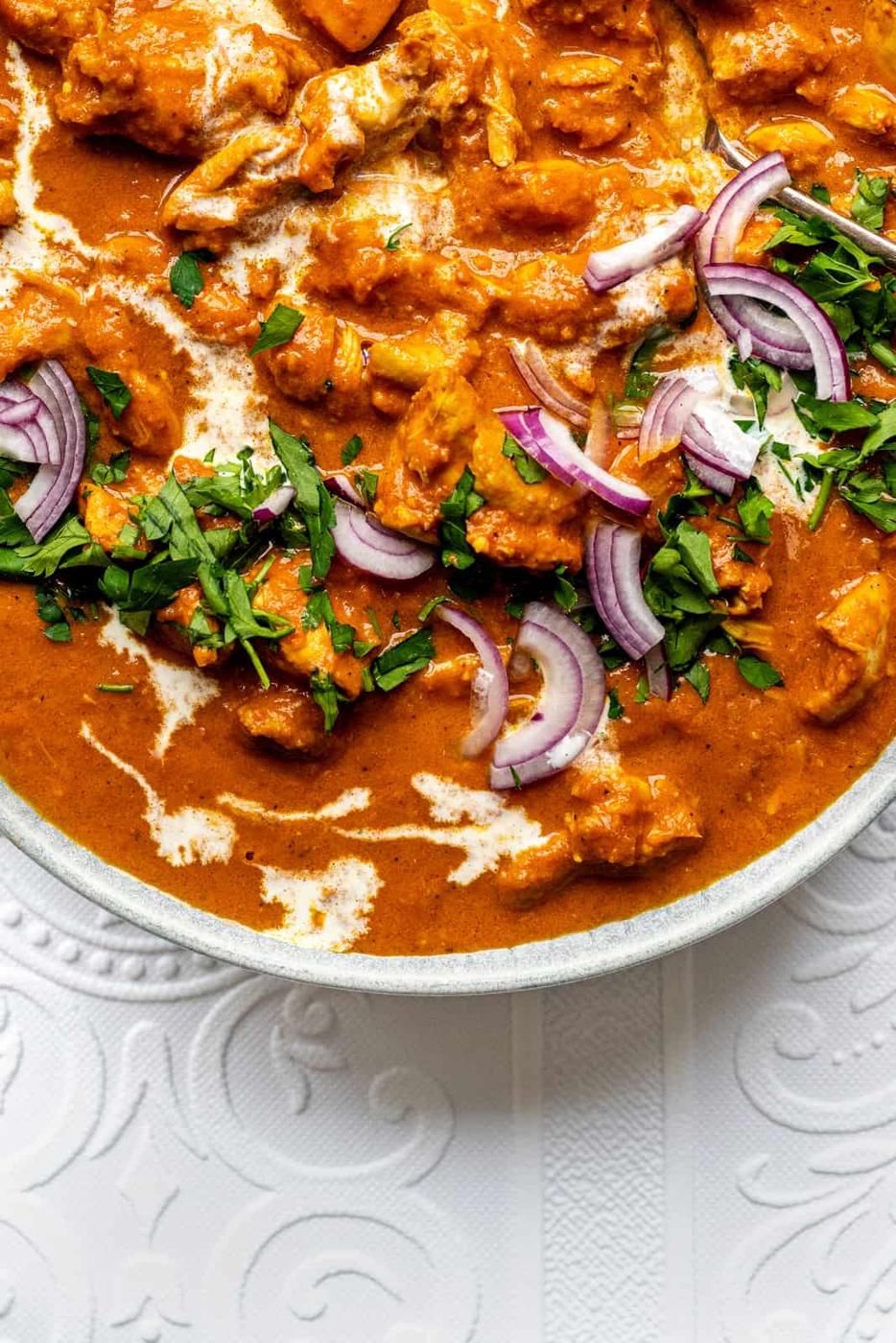 A bowl of creamy Butter Chicken curry garnished with fresh cilantro and sliced red onions, placed on a white embossed surface.