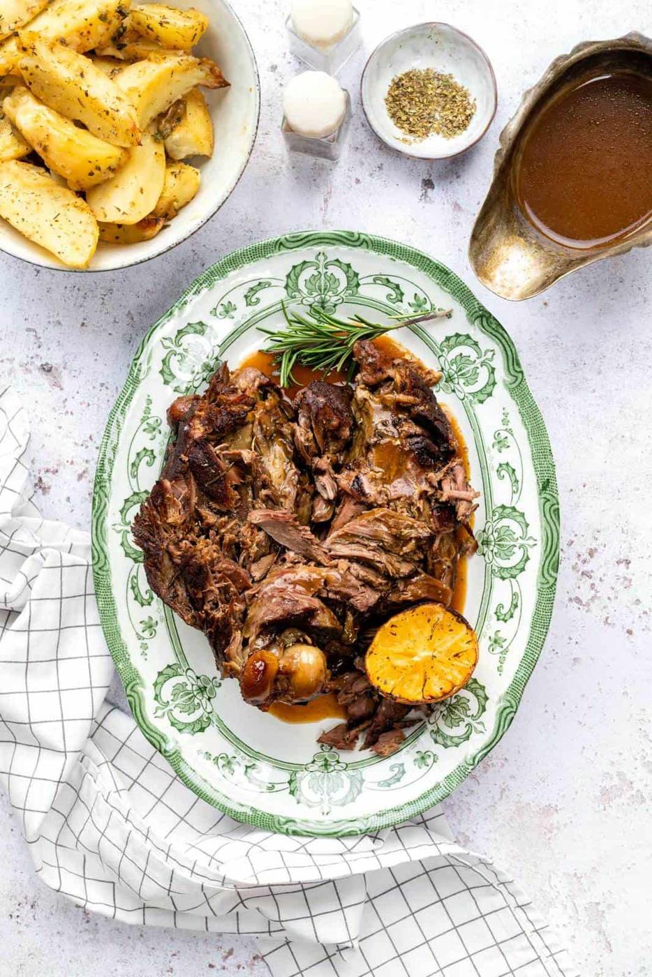 A green-rimmed platter with shredded Slow Cooker Lamb, roasted lemon, and rosemary sprigs, surrounded by a bowl of herbed potato wedges, gravy boat, seasonings, and a white checkered cloth.