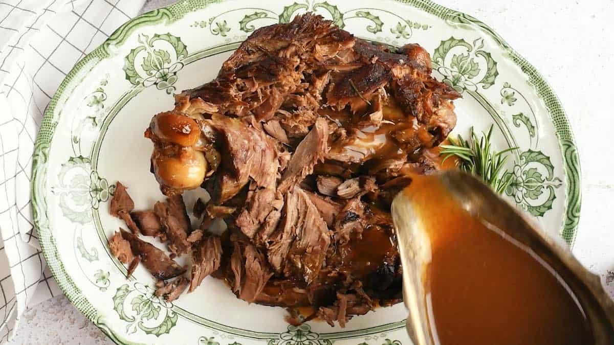 A platter of Slow Cooker Lamb, shredded with the bone, is served on a decorative green and white plate as brown gravy is poured over the tender meat. A sprig of rosemary sits on the side for garnish.