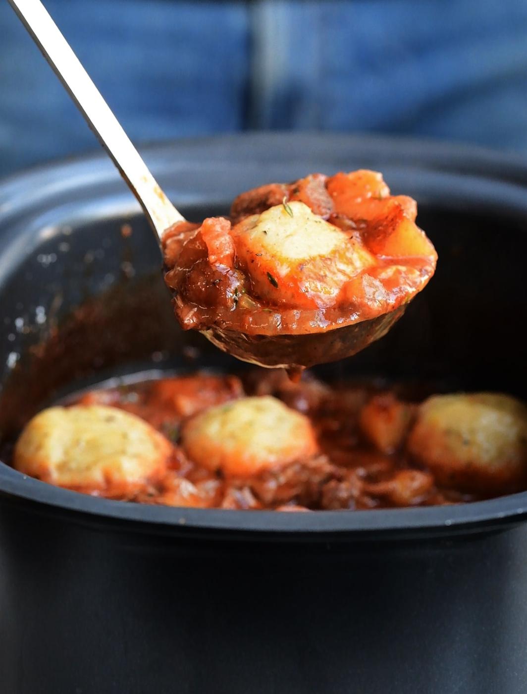 A close-up of a ladle lifting a serving of hearty lamb casserole in slow cooker, with chunks of vegetables and dumplings from a black crock pot. The rich, red sauce and visible herbs make this dish irresistible.
