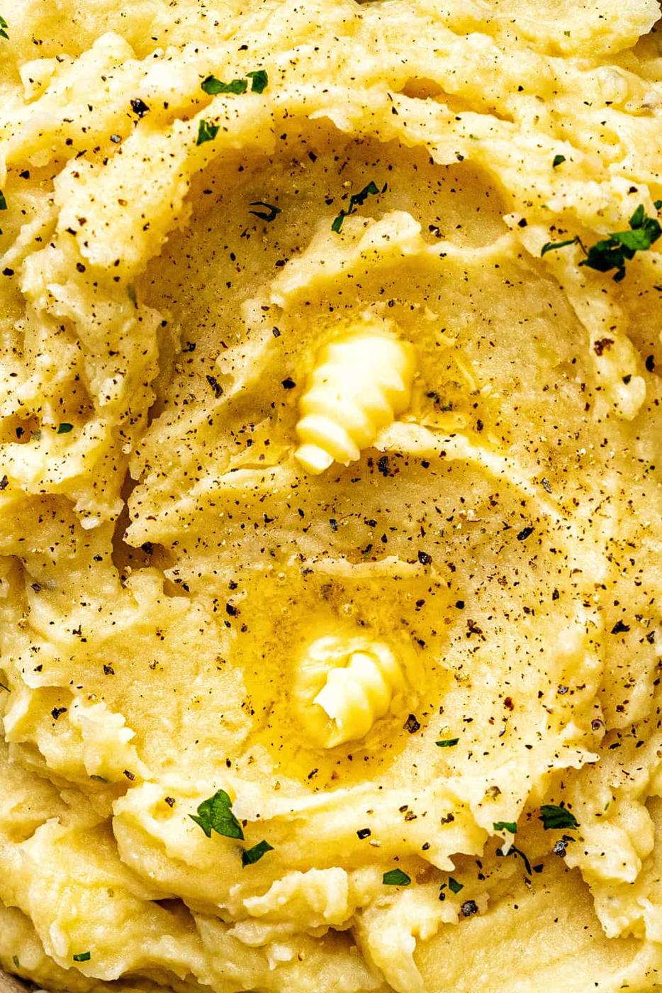 Close-up of creamy crockpot mashed potatoes topped with two melting pats of butter, freshly ground black pepper, and a sprinkle of chopped parsley.