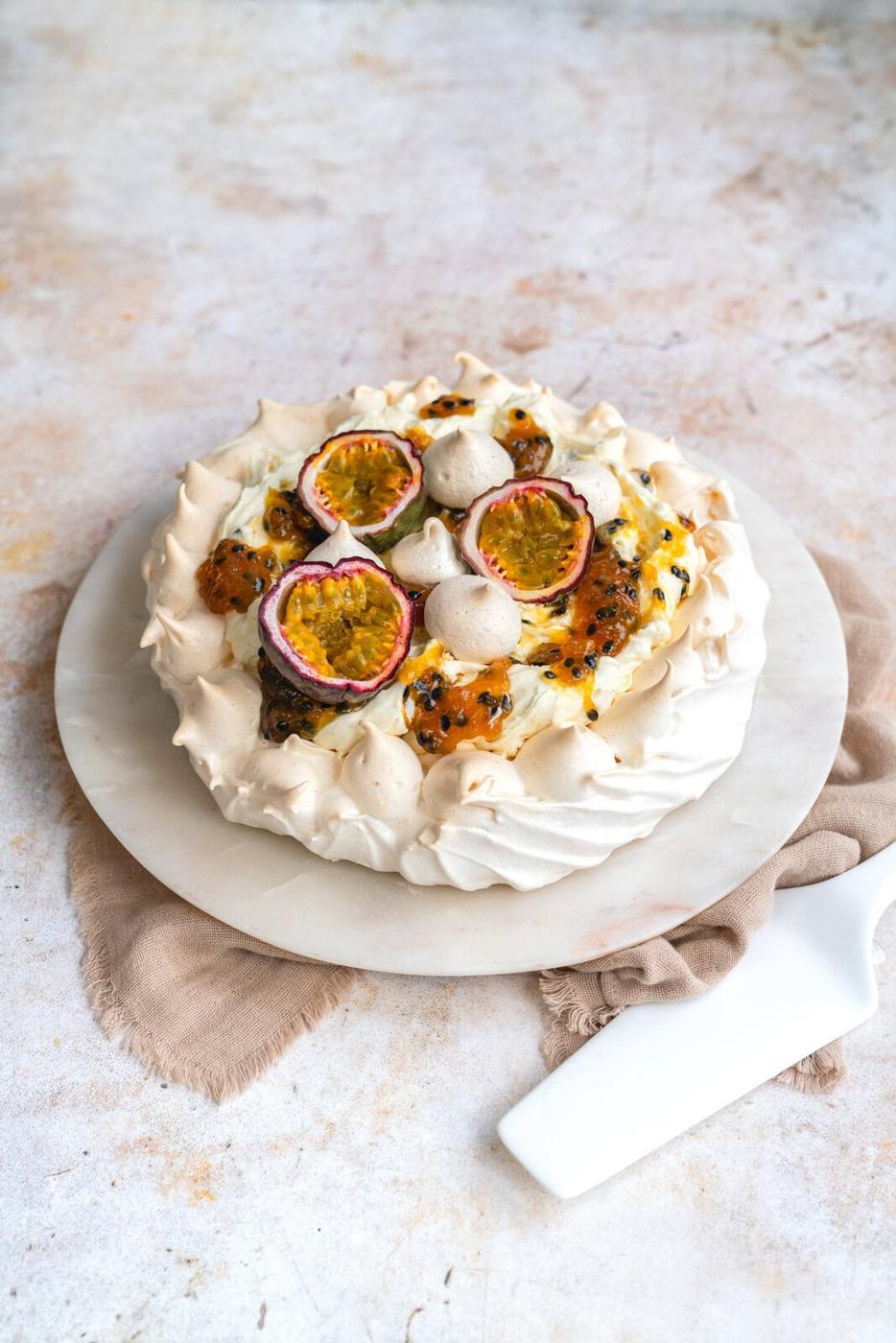 A pavlova topped with passion fruit halves, meringue kisses, and cream is placed on a white marble platter. A white cake server lies beside it on a beige cloth over a textured surface.