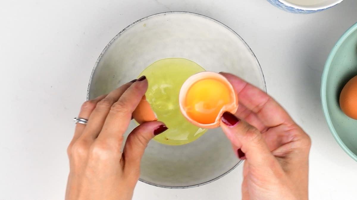 Hands are cracking a brown egg over a white bowl, revealing the yolk and white—perfect for crafting a delicate Black Forest Pavlova. Nearby, another egg rests in a light blue dish on the light-colored surface, waiting to join in the culinary creation.