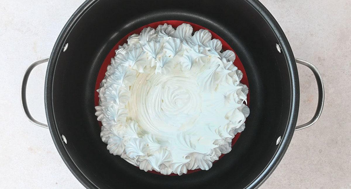 Top view of a meringue in a circular pattern, sitting inside a slow cooker insert. The meringue has a swirled texture and is placed on a red base within the pan. The pan is on a light-colored surface.