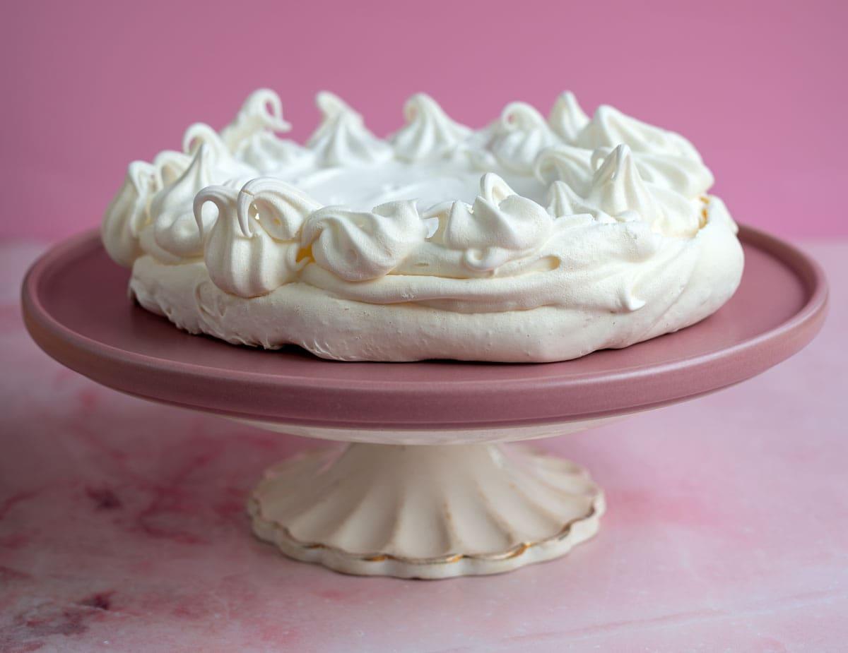 A round pavlova with fluffy white meringue peaks is placed on a pink cake stand. The background is a pastel pink