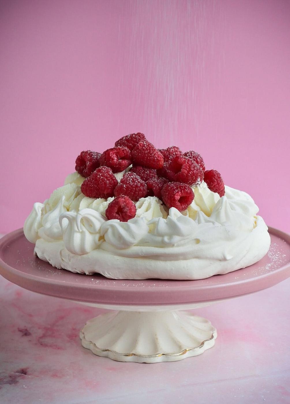 A pavlova topped with fresh raspberries sits on a pink cake stand. The background is pink, and powdered sugar is being sprinkled over the dessert.
