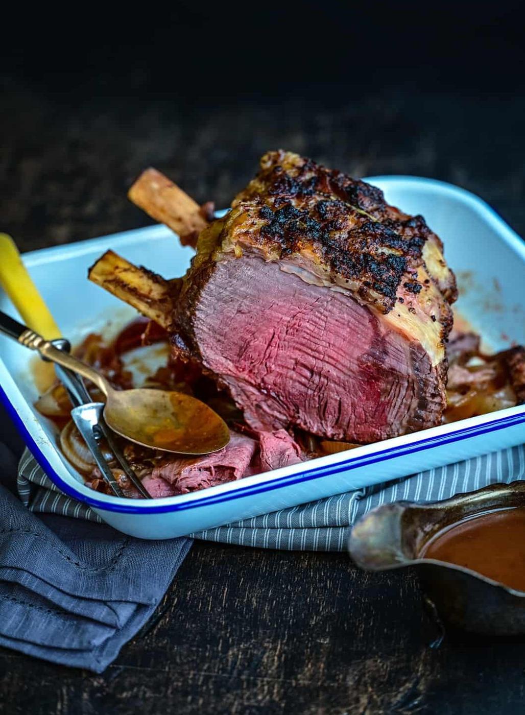 A Slow Cooker Prime Rib with a crispy, seasoned crust and pink center is partially sliced in a white roasting pan. Slices and utensils are nearby, with a serving of brown sauce in a small gravy boat beside the pan.