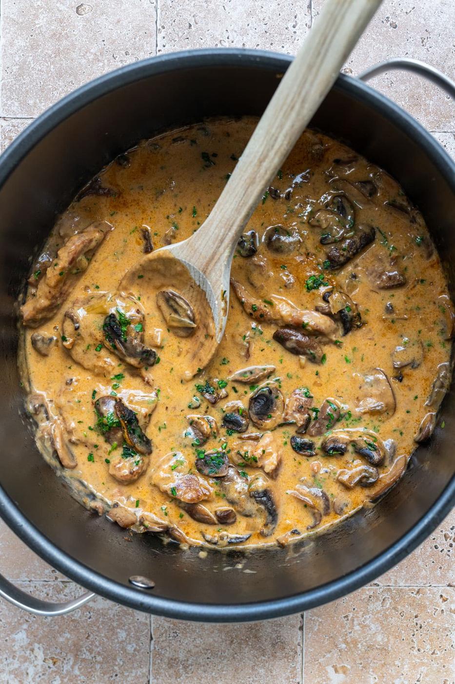 Crock pot insert with pork chops in a creamy mushroom sauce with sliced mushrooms and herbs, stirred with a wooden spoon. The sauce is rich and thick, covering the ingredients. The pot sits on a light marbled surface.