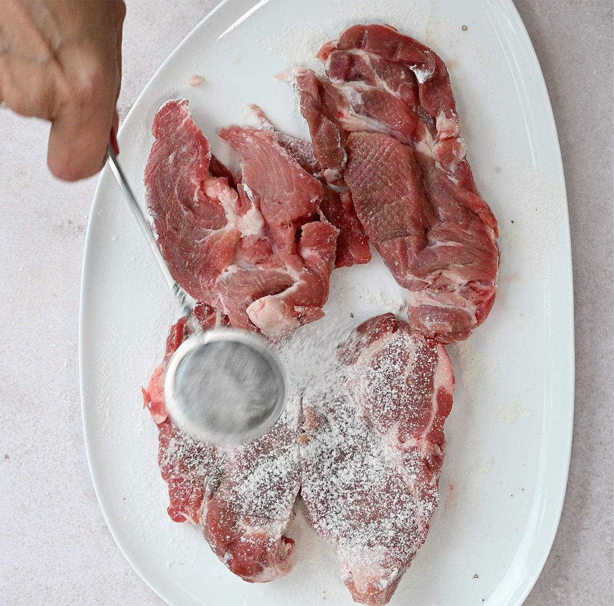 A hand sprinkles salt over raw pork steaks arranged on a white oval plate. The background is a light-colored surface.