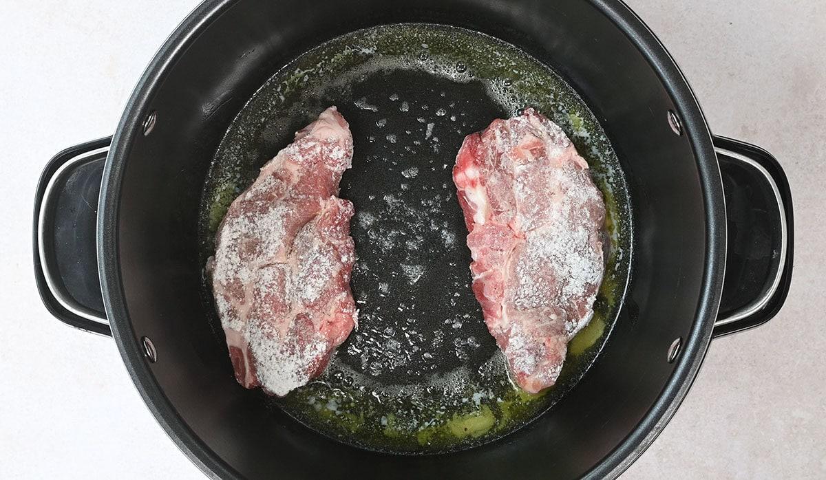 Two raw steaks in a large black pot, partially coated with seasoning, are beginning to sizzle in melted butter on a stovetop. The pot has two handles, and the steaks are placed side by side.