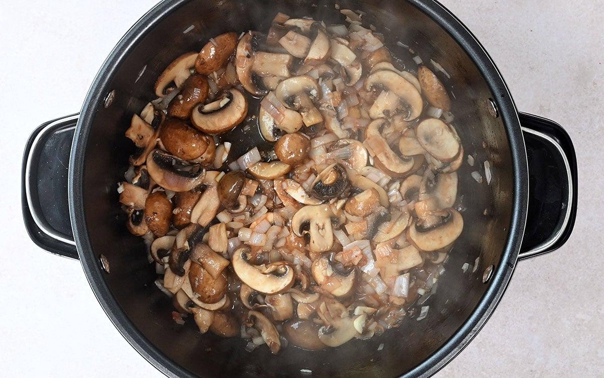 A pot filled with sliced mushrooms and diced onions being sautéed. The mushrooms are a mix of brown and white varieties, and they are lightly browned, suggesting they are being cooked. Steam rises from the pot.