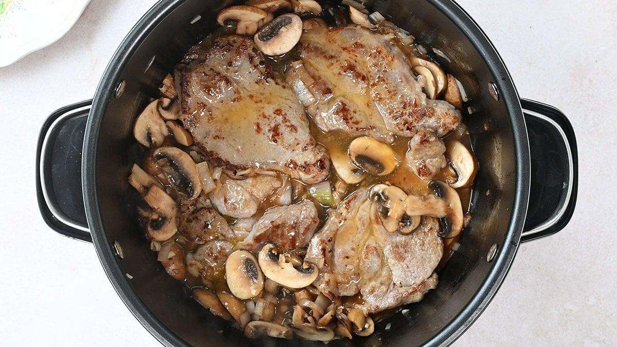 A slow cooker filled with pork steaks, sliced mushrooms, and onions in a brown sauce. The ingredients appear tender and well-coated with the sauce, creating a savory dish.