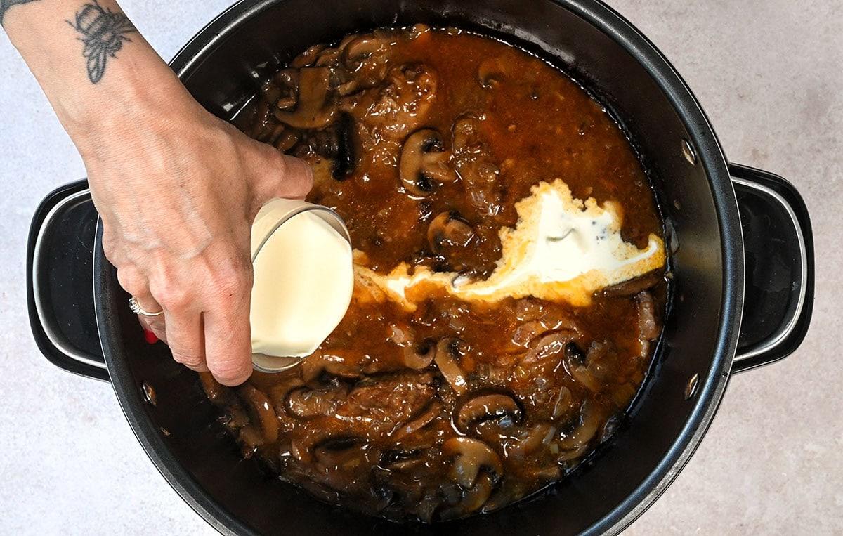 A hand with a tattoo is pouring cream into a pot of smothered pork steaks
