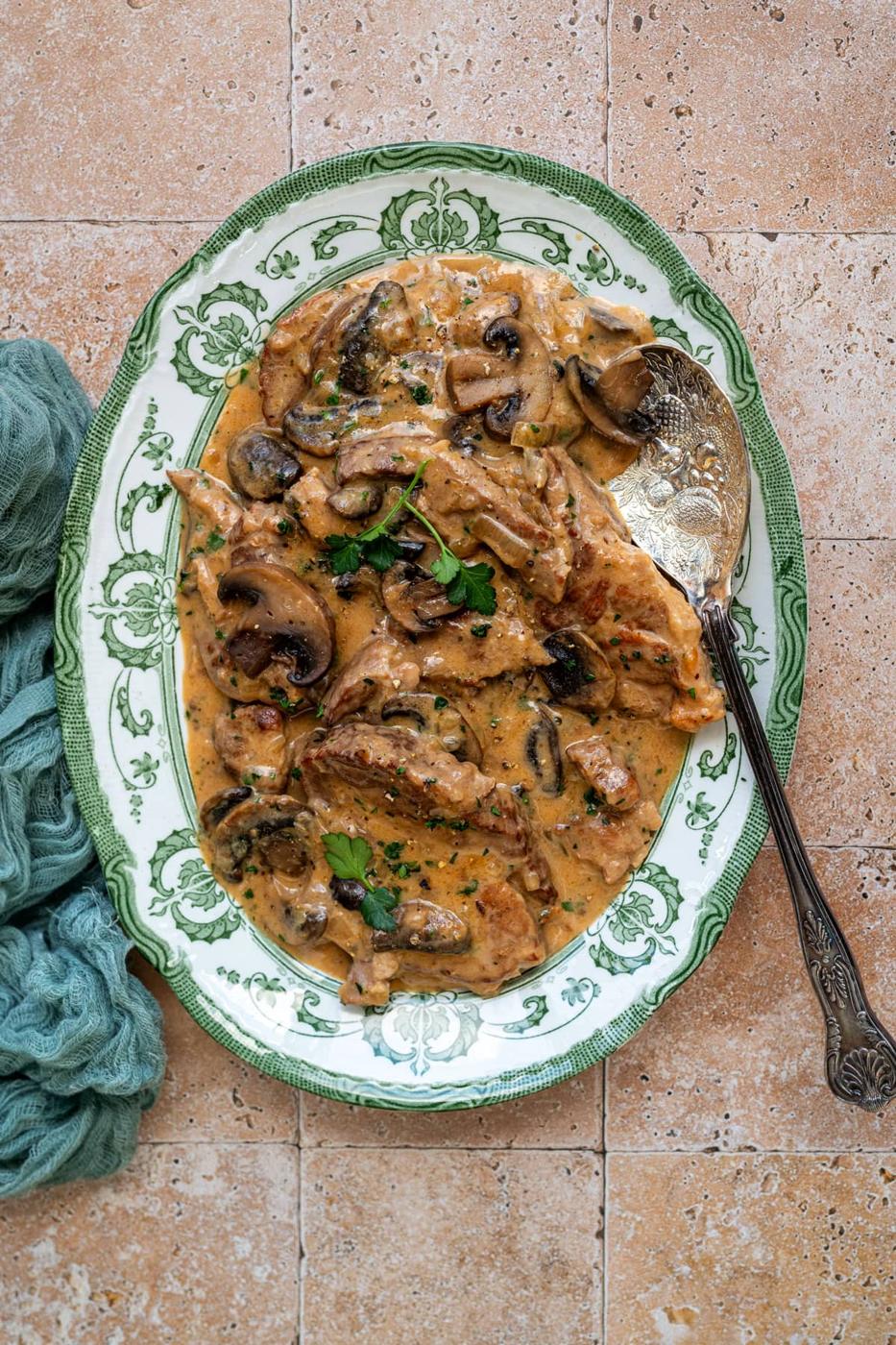 A decorative plate filled with pork marsala, featuring pork shoulder steaks topped with a creamy sauce, mushrooms, and garnished with parsley. A vintage silver spoon rests beside the dish on a tiled surface, accompanied by a green cloth.