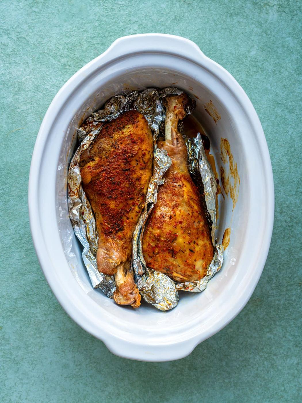 Two seasoned turkey legs wrapped in aluminum foil, placed inside a white oval slow cooker on a green surface.