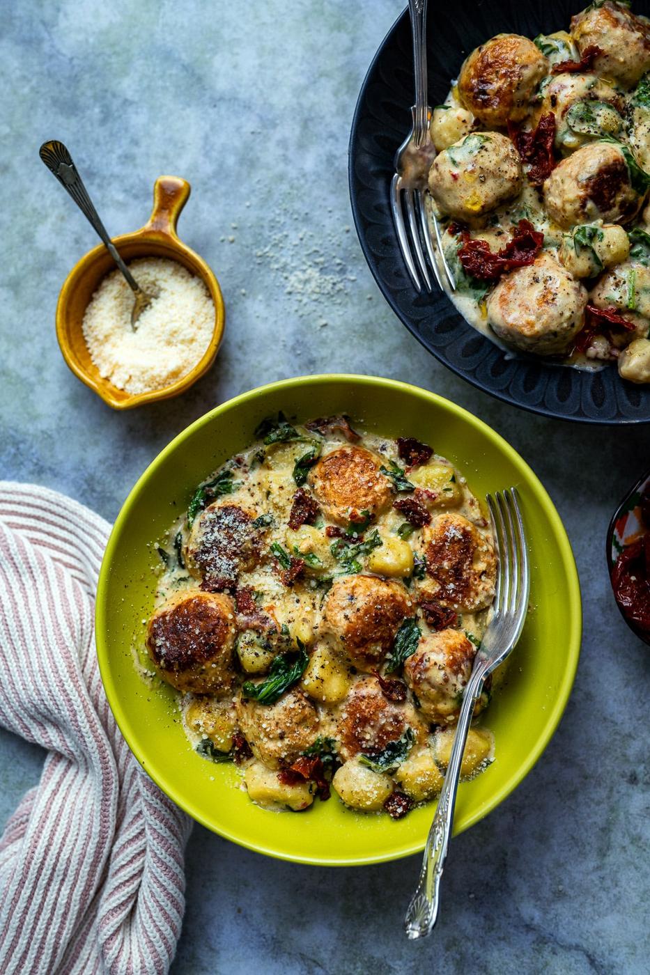 A yellow bowl filled with Slow Cooker Tuscan Chicken Meatballs with Gnocchi, creamy spinach, and sun-dried tomatoes, garnished with grated cheese. A fork rests in the bowl. A small dish of grated cheese and a striped napkin are nearby.