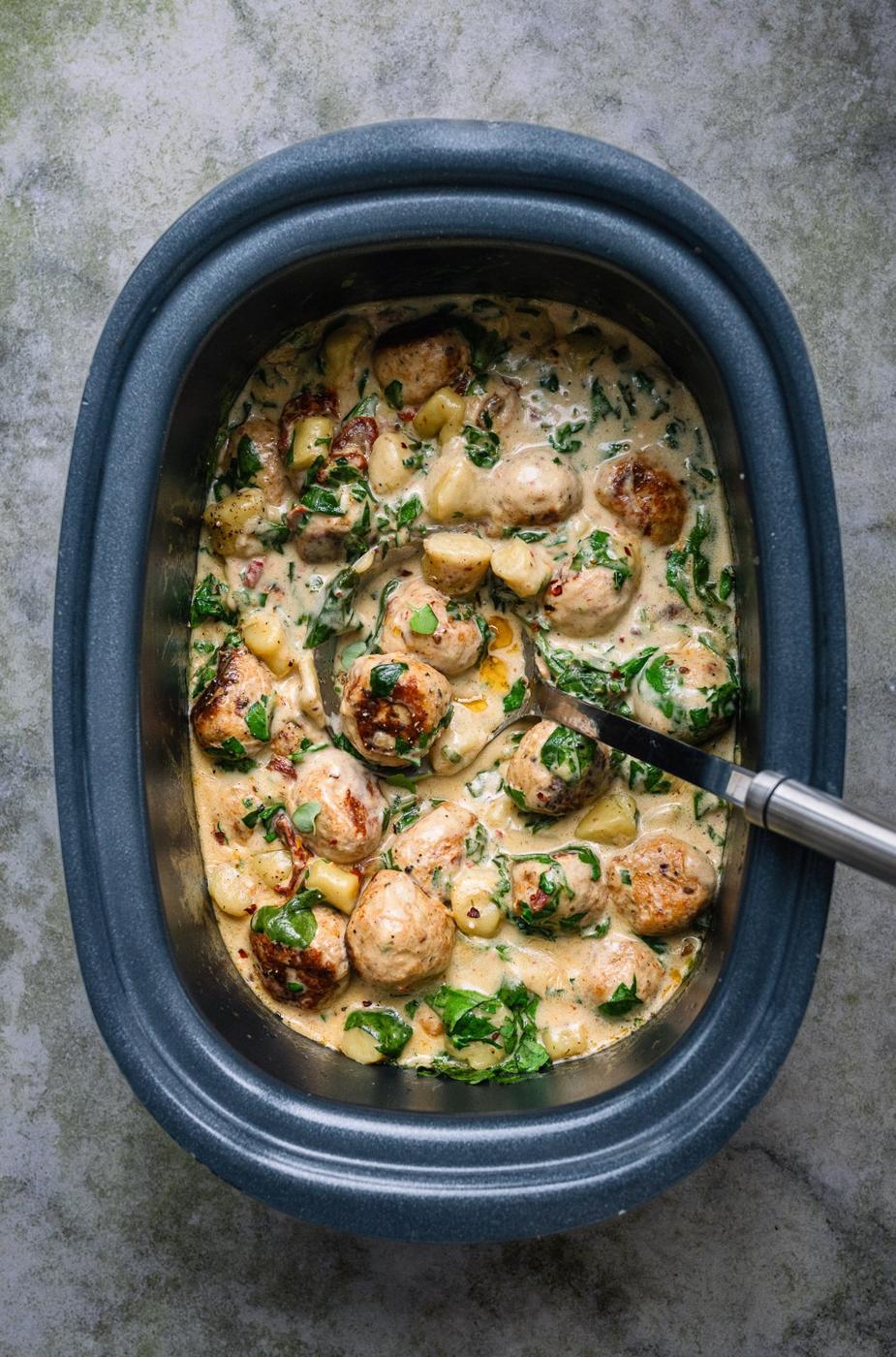 A slow cooker filled with creamy sauce, golden-brown Slow Cooker Tuscan Chicken Meatballs with Gnocchi, and spinach, with a serving spoon resting inside.