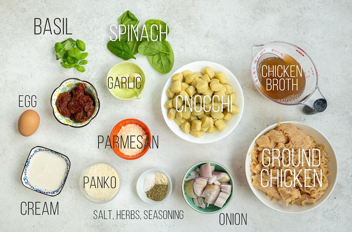 Top-down view of labeled ingredients for Slow Cooker Tuscan Chicken Meatballs with Gnocchi, including basil, spinach, garlic, chicken broth, ground chicken, gnocchi, onion, herbs, panko, parmesan, cream, egg, and sun-dried tomatoes.