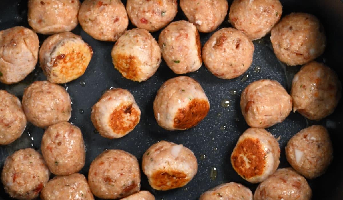 A close-up view of several Slow Cooker Tuscan Chicken Meatballs with Gnocchi sizzling in a pan, some with browned sides, arranged closely together with a slight sheen of oil visible.