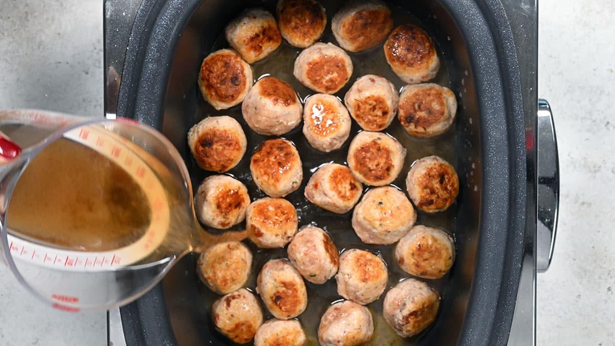 A slow cooker filled with Slow Cooker Tuscan Chicken Meatballs with Gnocchi is shown from above. A hand pours broth or sauce from a measuring cup over the savory meatballs.