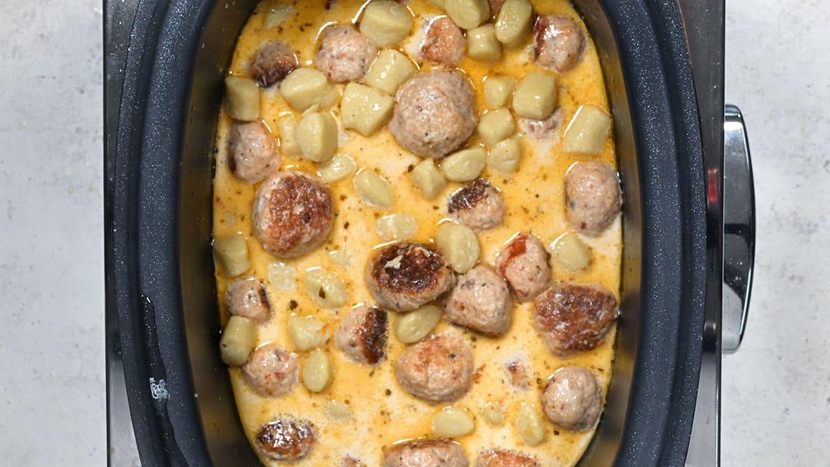 A slow cooker filled with Slow Cooker Tuscan Chicken Meatballs with Gnocchi, creamy sauce, browned meatballs, and chunks of potatoes, viewed from above.