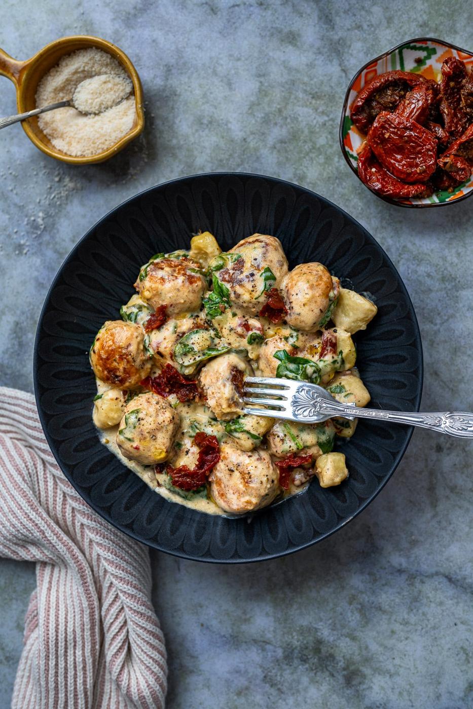 A black bowl filled with creamy chicken, gnocchi, sun-dried tomatoes, and spinach – reminiscent of Slow Cooker Tuscan Chicken Meatballs with Gnocchi. A silver fork rests in the bowl beside a striped napkin and a small bowl of grated cheese.