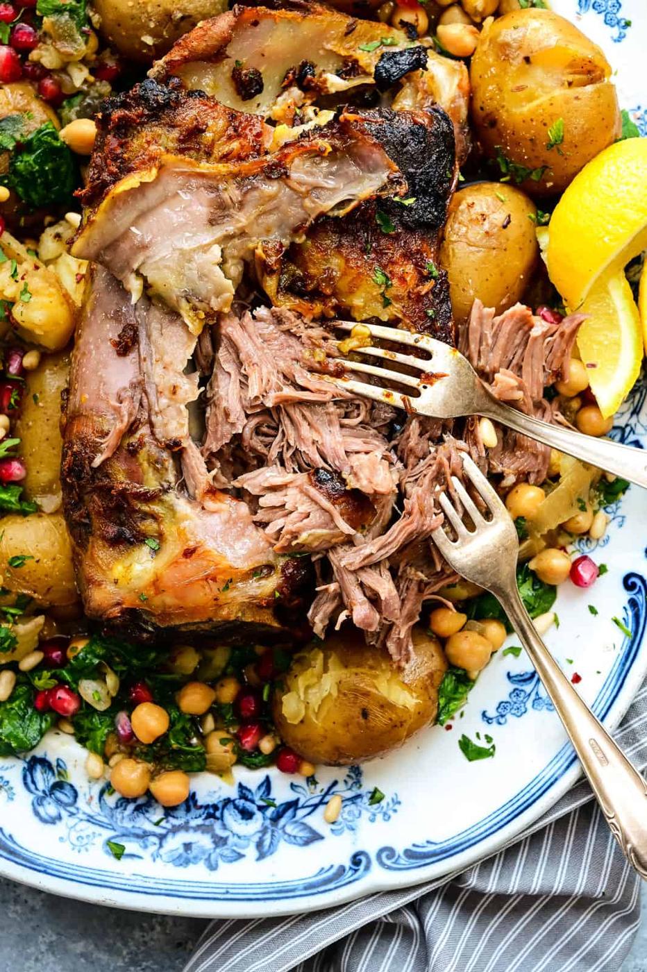 Close up on tender slow roasted lamb shoulder on a bed of potatoes, chickpeas and spinach, being shredded with two forks
