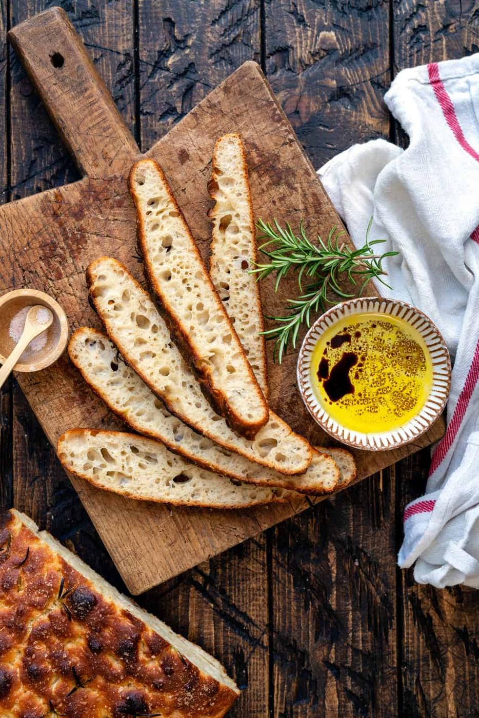 Sliced sourdough focaccia on a wooden board with rosemary, olive oil and balsamic vinegar, salt, and a white towel with red stripes—a perfect setup for anyone following an easy sourdough focaccia recipe.