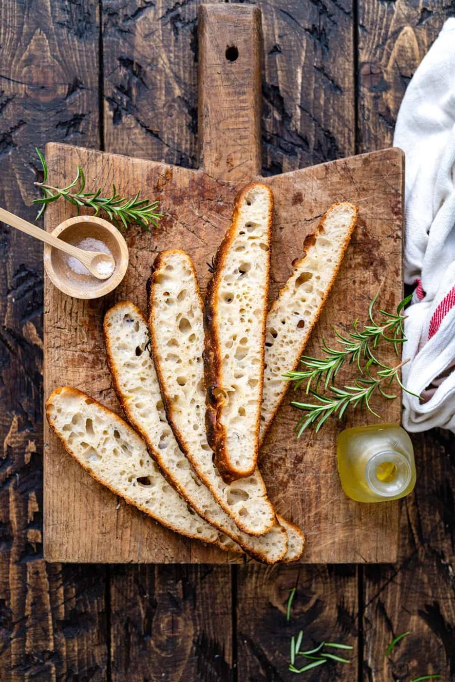 Four slices of rustic sourdough bread on a wooden cutting board with rosemary, a bowl of salt, and olive oil invite you to try an easy sourdough focaccia recipe—the perfect start for beginners seeking delicious homemade bread.