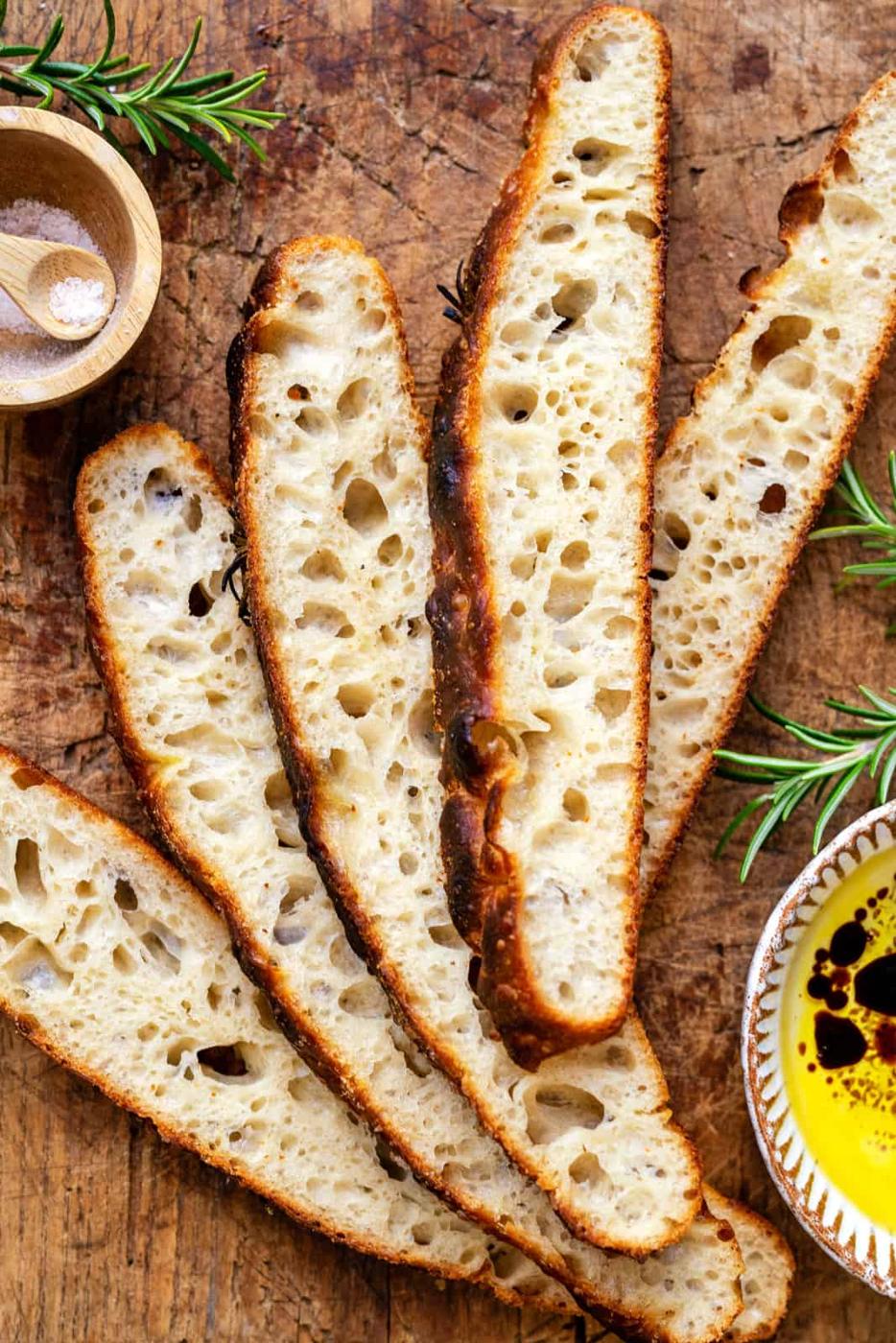 Five slices of rustic, airy sourdough focaccia rest on a wooden surface, surrounded by rosemary sprigs, a small bowl of salt, and a dish of olive oil and balsamic vinegar—perfect for anyone seeking an easy sourdough focaccia recipe.