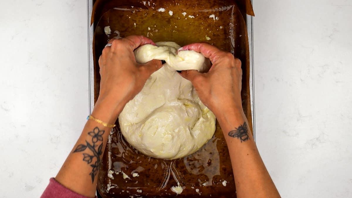 A person with floral tattoos on their forearms is stretching dough in a parchment-lined tray, preparing an easy sourdough focaccia recipe.